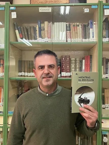 El autor, vestido de verde oliva, posa con su libro Mientras el río fluye, de fondo verde oliva, delante de una estantería también verde oliva.