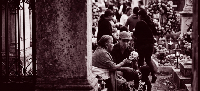 Elderly woman, man, and child in skeleton costume interacting with a skull in a cemetery during Day of the Dead