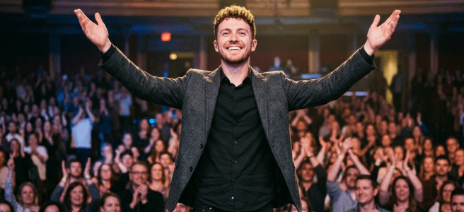 Man standing on stage with arms raised while audience applauds