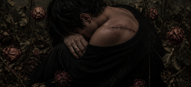 Wounded person sitting among withered roses; text reads “HIJO Y HERIDA” and “ENTRE ROSAS MARCHITAS”